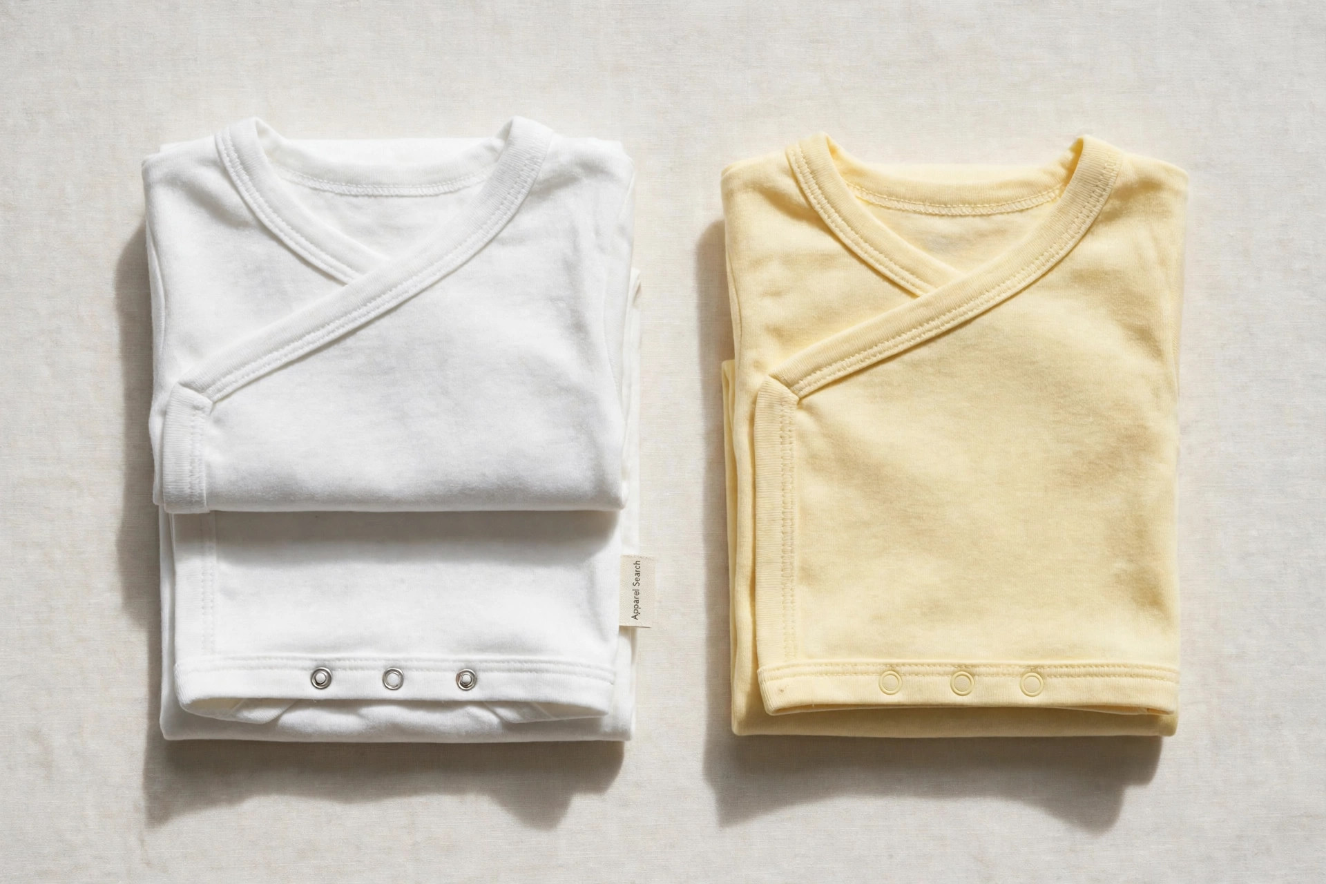 Folded baby bodysuits in soft white and pale yellow cotton laid flat, wrap necklines and bottom snaps visible with a small printed Apparel Search label, product photography with no people
