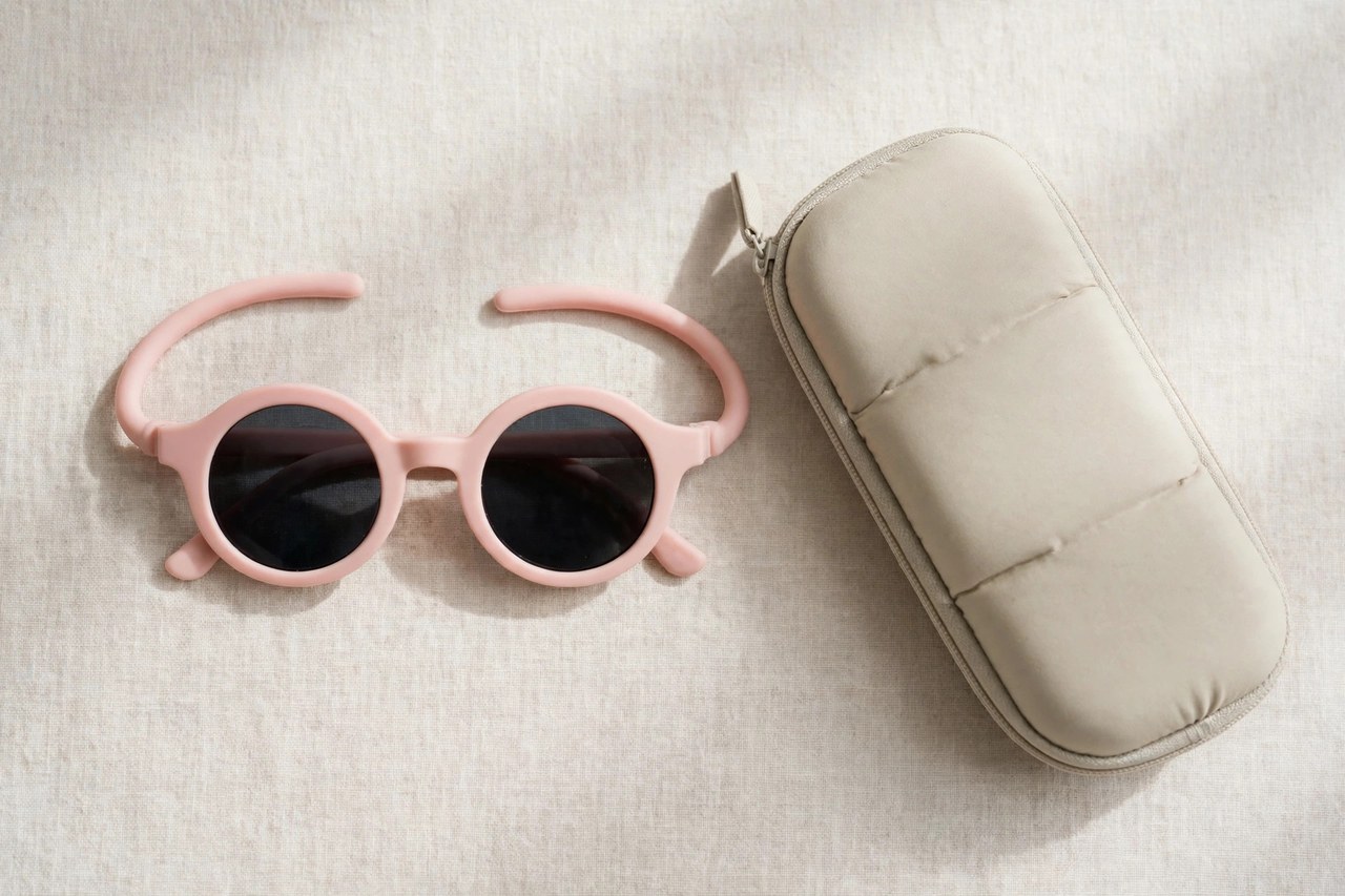 A flat lay of children's sunglasses with flexible frames and dark UV-protective lenses beside a soft case, arranged on a clean background, product photography with no people, no children and no mannequins