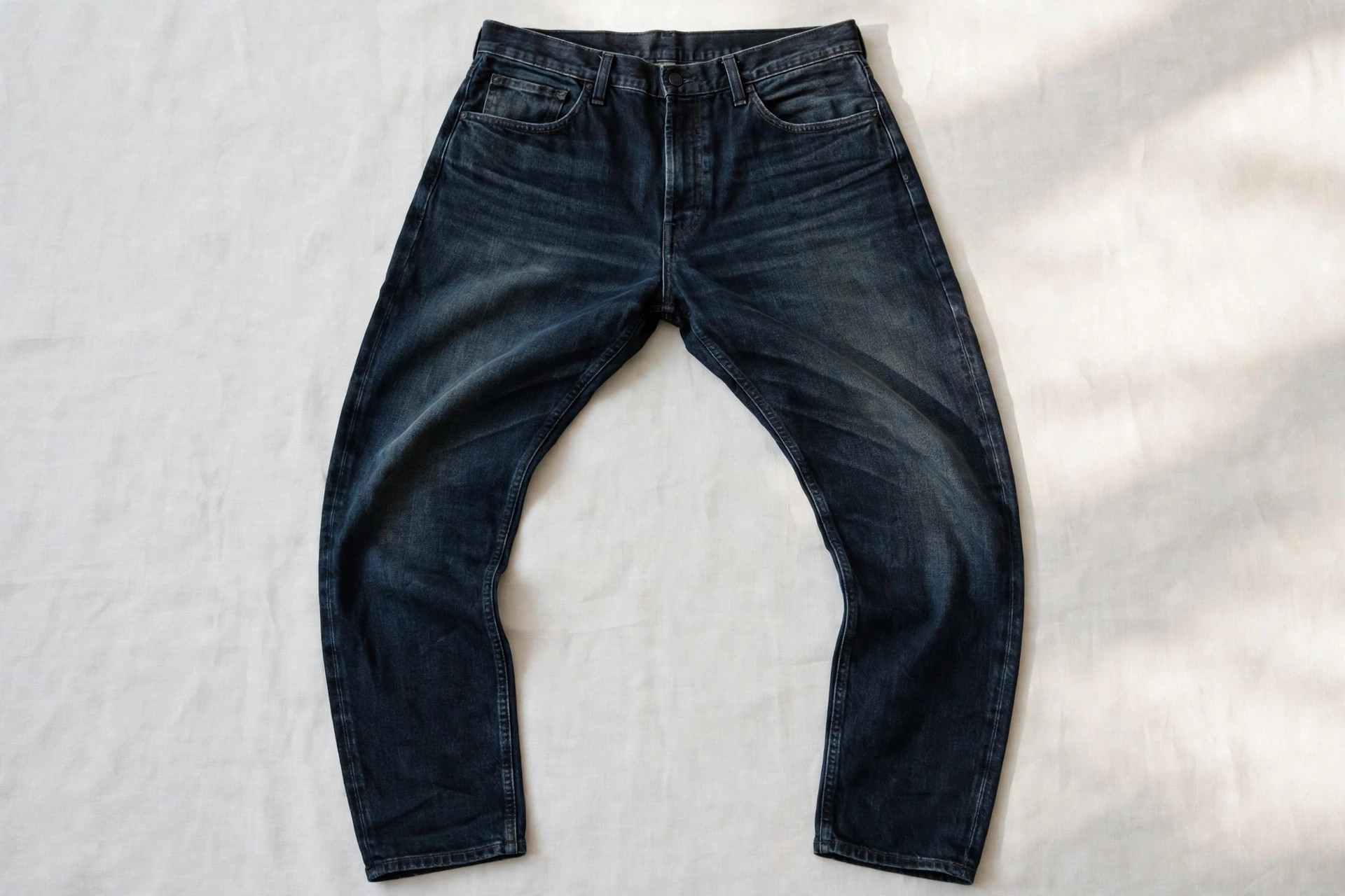 Men's barrel jeans in a dark washed denim laid flat, showing the curved leg bowing out at the thigh and tapering to a clean ankle, product photography with no people