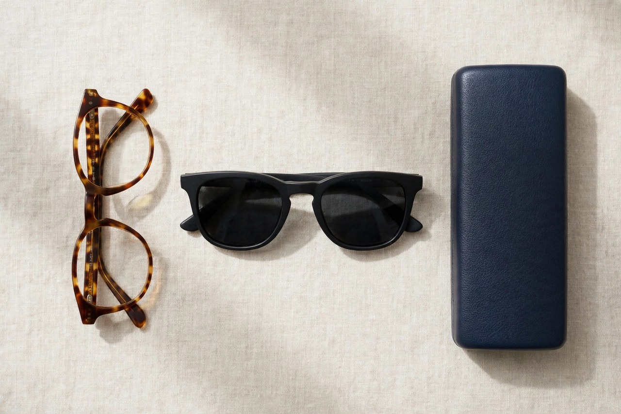 A flat lay of men's eyewear: a pair of tortoiseshell eyeglasses, dark sunglasses and a hard case arranged neatly, product photography with no people