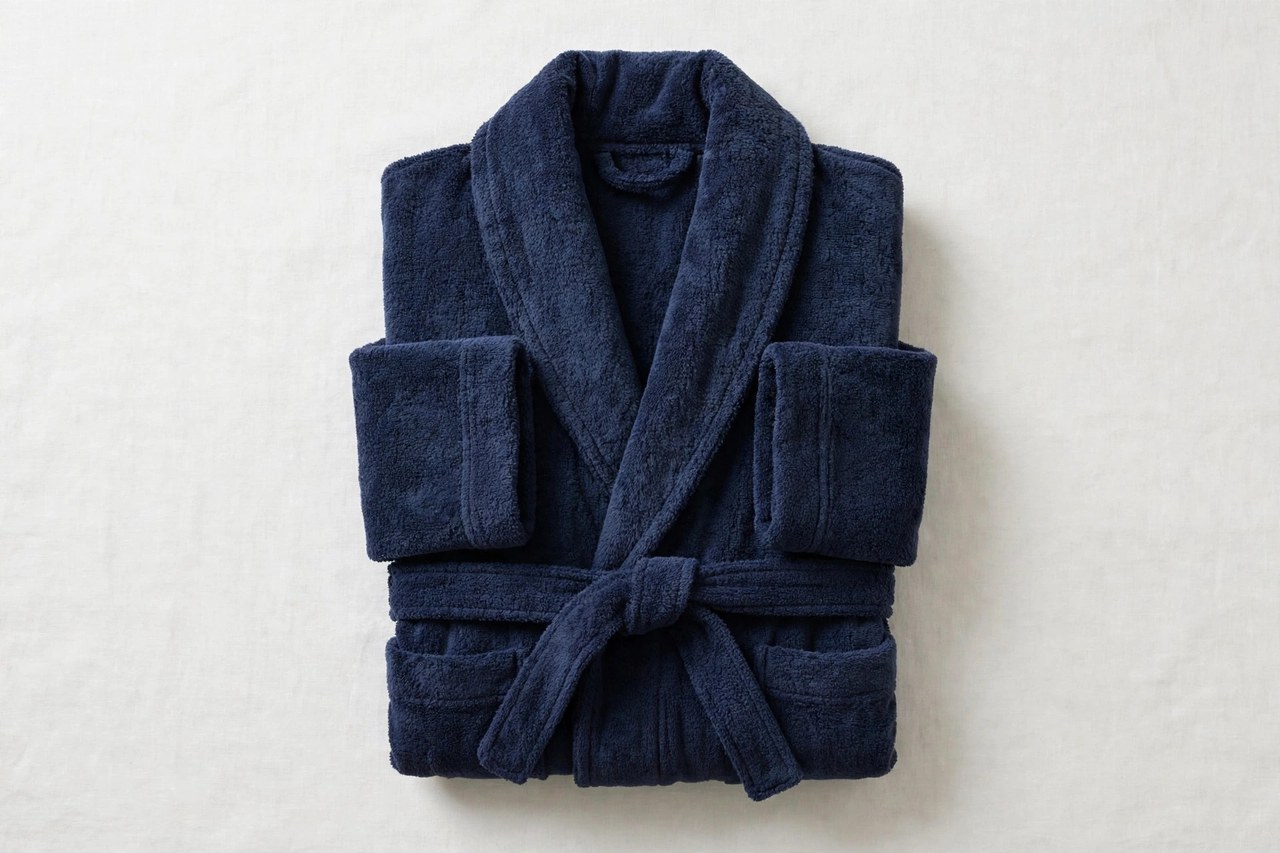 A flat lay of a men's plush terry cloth bathrobe in deep navy with a shawl collar and tie belt, folded neatly, product photography with no people