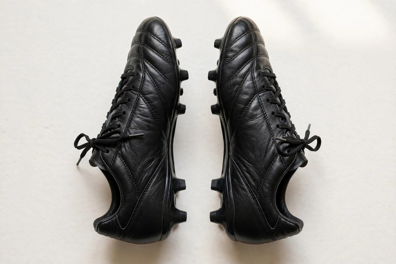 A flat lay of a pair of men's soccer cleats in black leather with molded studs, arranged on a clean background, product photography with no people