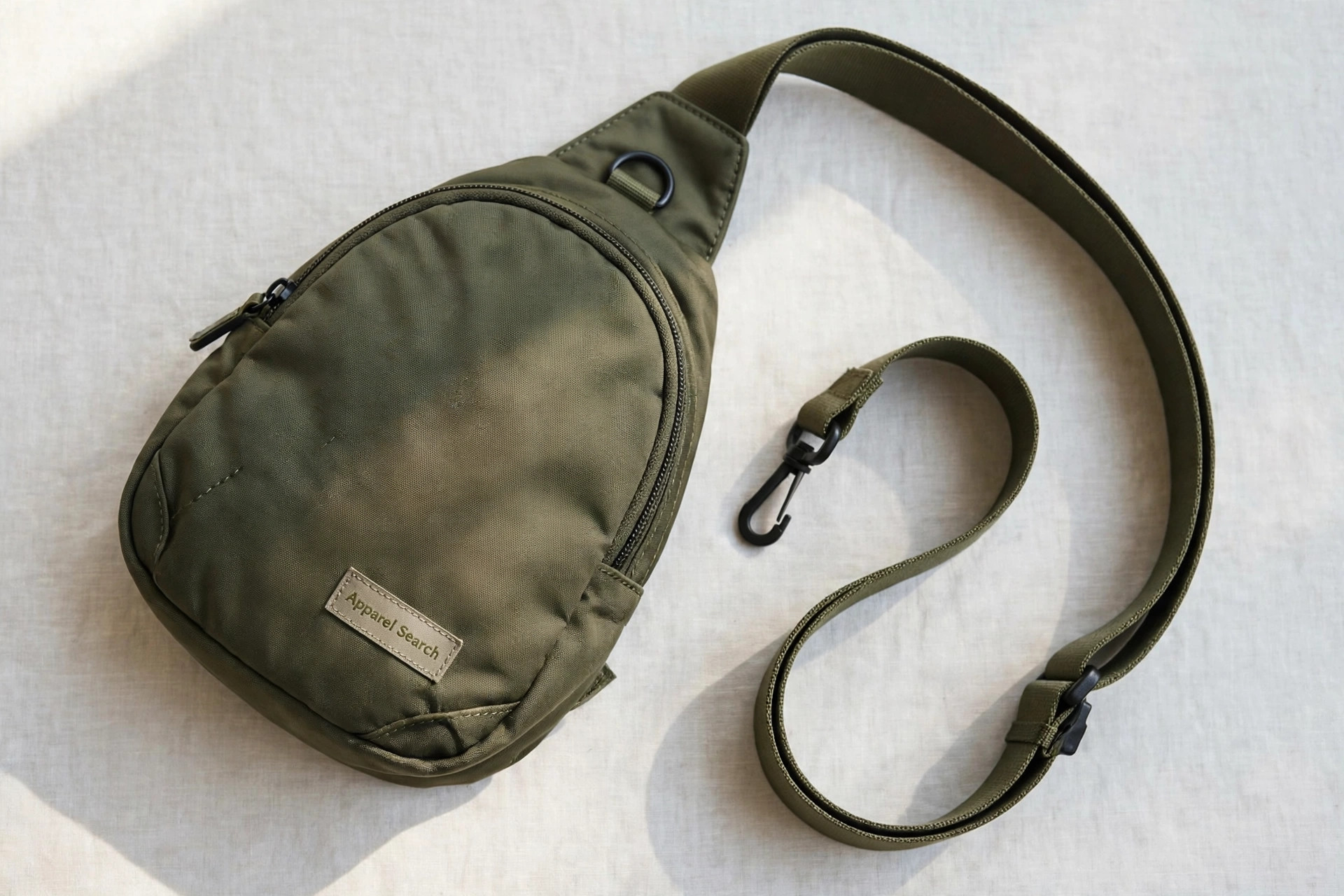 Women's sling bag in olive nylon with a wide adjustable strap laid curved beside it, product photography with no people