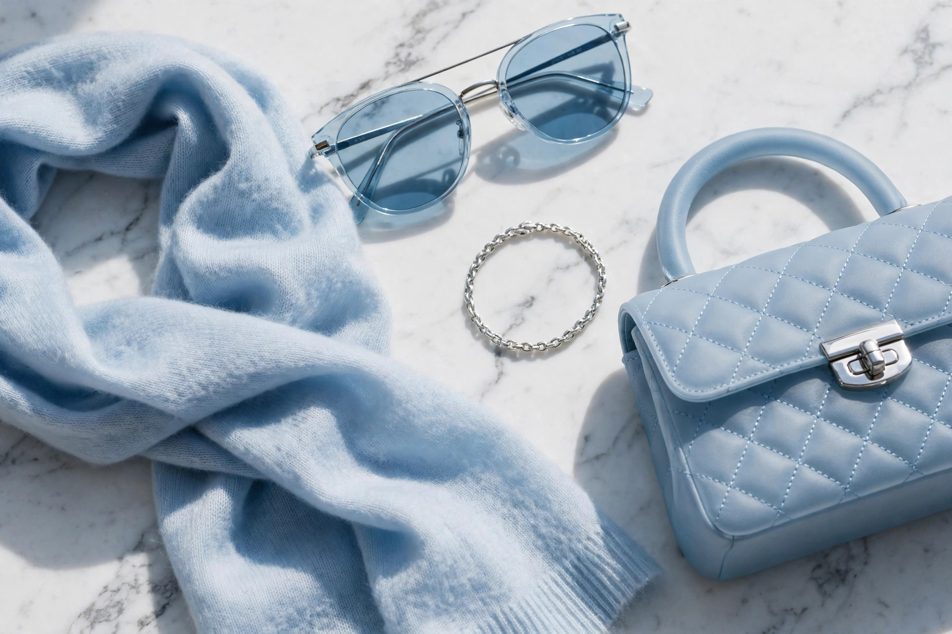 Pale blue cashmere scarf, light blue tinted sunglasses, a silver bracelet and a frosty blue handbag arranged on white marble in cool daylight