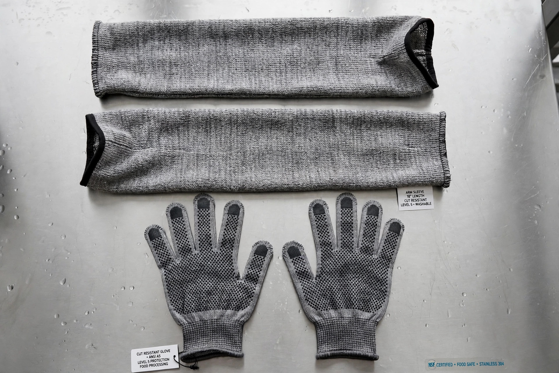 Gray knitted cut-resistant arm sleeves and cut-resistant gloves arranged on a stainless steel surface