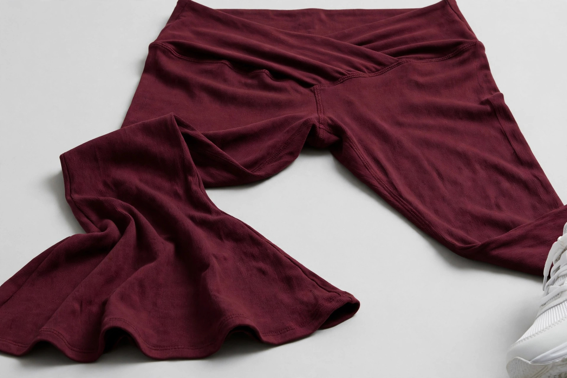 Close-up of the flared hem and crossover waistband of flare leggings in deep burgundy laid flat on a studio background with sneakers nearby