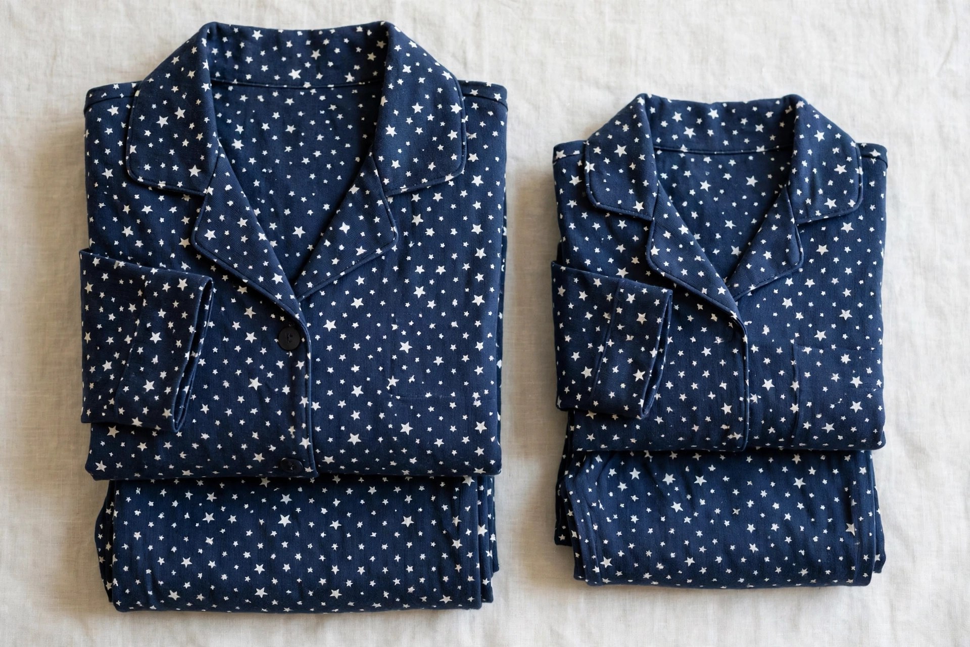 Flat lay of matching pajama sets in soft navy cotton with a white star print, adult and smaller sizes folded side by side, garments only